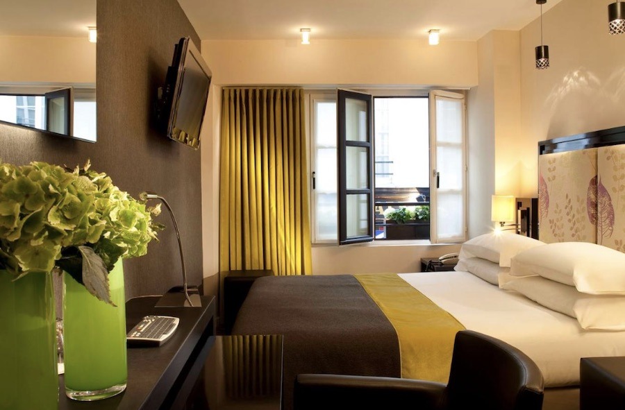This is an image of a hotel room with a large double bed in the centre and lots of pillows. The room also has green curtains that are half open.