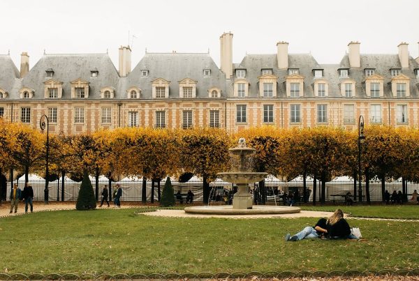 Row of Parisian townhouses with dormer windows overlooking Place des Vosges in autumn, with golden-leaved trees, a stone fountain, and two people relaxing on the grass.