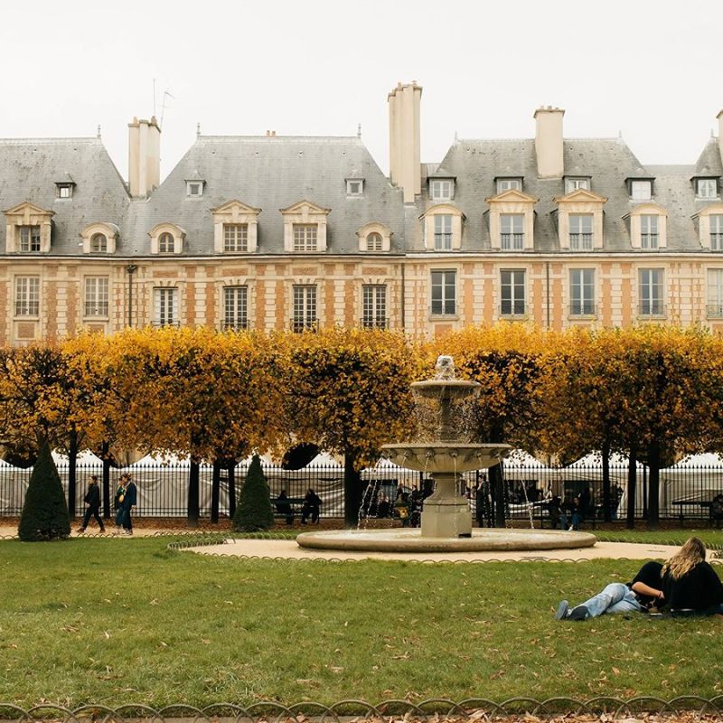 Row of Parisian townhouses with dormer windows overlooking Place des Vosges in autumn, with golden-leaved trees, a stone fountain, and two people relaxing on the grass.