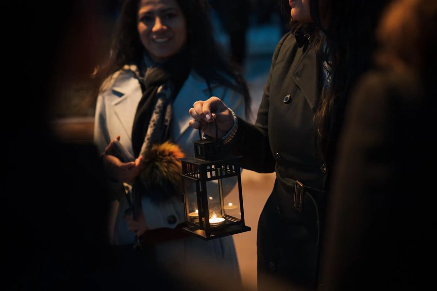 Nighttime ghost tour scene showing a woman holding a lit lantern, illuminating her face and a smiling companion beside her in a dark, atmospheric setting.