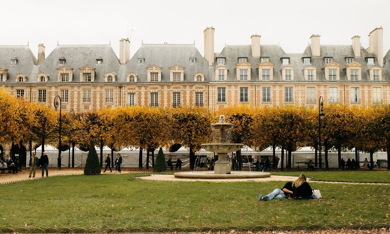Row of Parisian townhouses with dormer windows overlooking Place des Vosges in autumn, with golden-leaved trees, a stone fountain, and two people relaxing on the grass.
