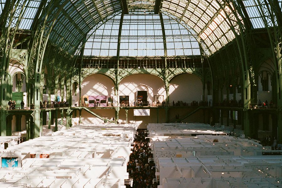Interior view of the Grand Palais in Paris during an art fair, showing rows of white exhibition booths beneath the building's glass-and-iron domed roof, with visitors browsing below.