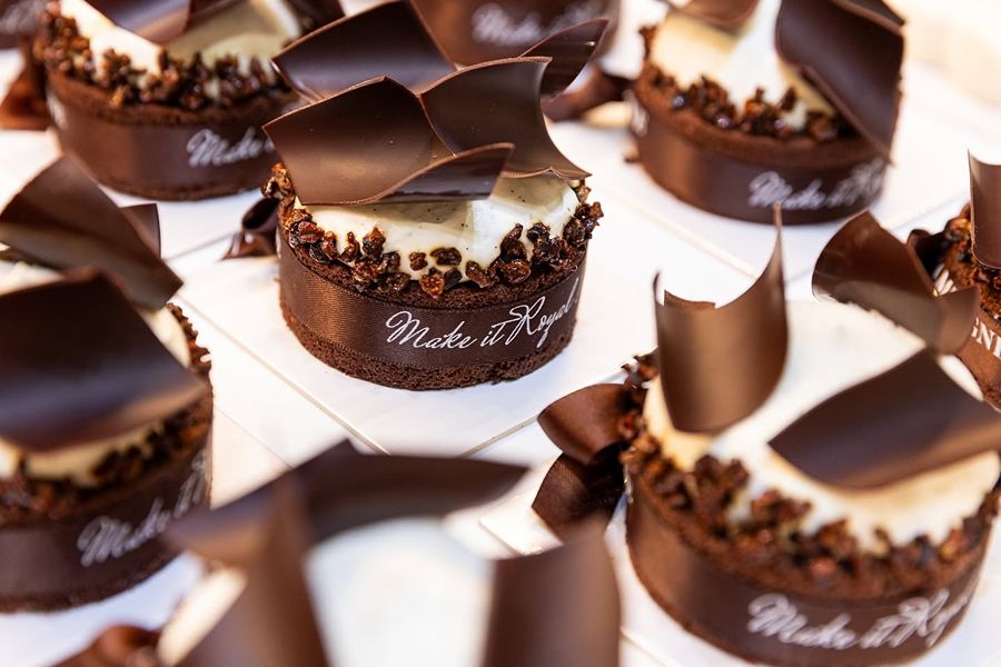 Close-up of individual round chocolate tarts topped with vanilla cream, cocoa nib crumble, and curled dark chocolate shards, each wrapped in a brown ribbon reading 'Make it Royal