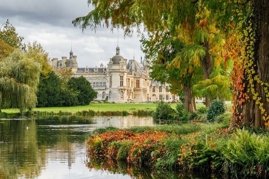 Château de Chantilly seen across a calm lake, framed by autumn foliage and a large tree draped in orange ivy, with turrets and a domed tower reflected in the water.