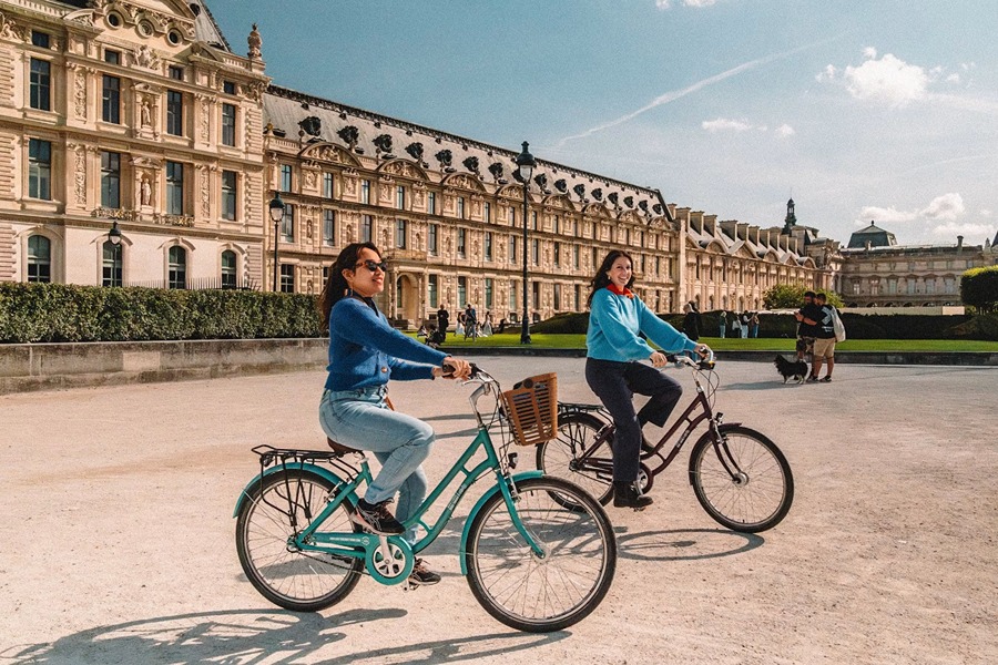 Two women cycling past the Louvre on a bike tour, one of the best things to do in September in Paris.