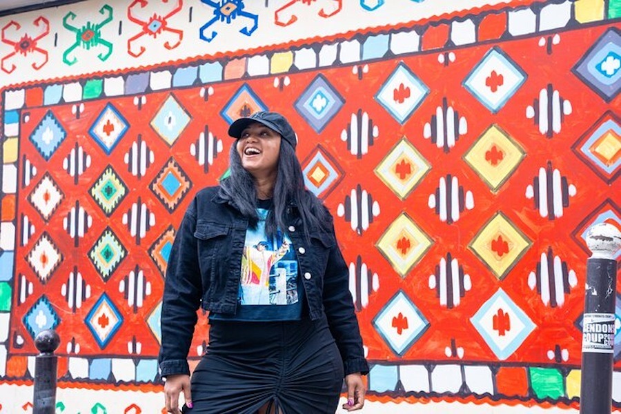 This is an image of a woman stood in front of a vibrant red background. She is wearing cool casual wearing that is dark and looks happy.