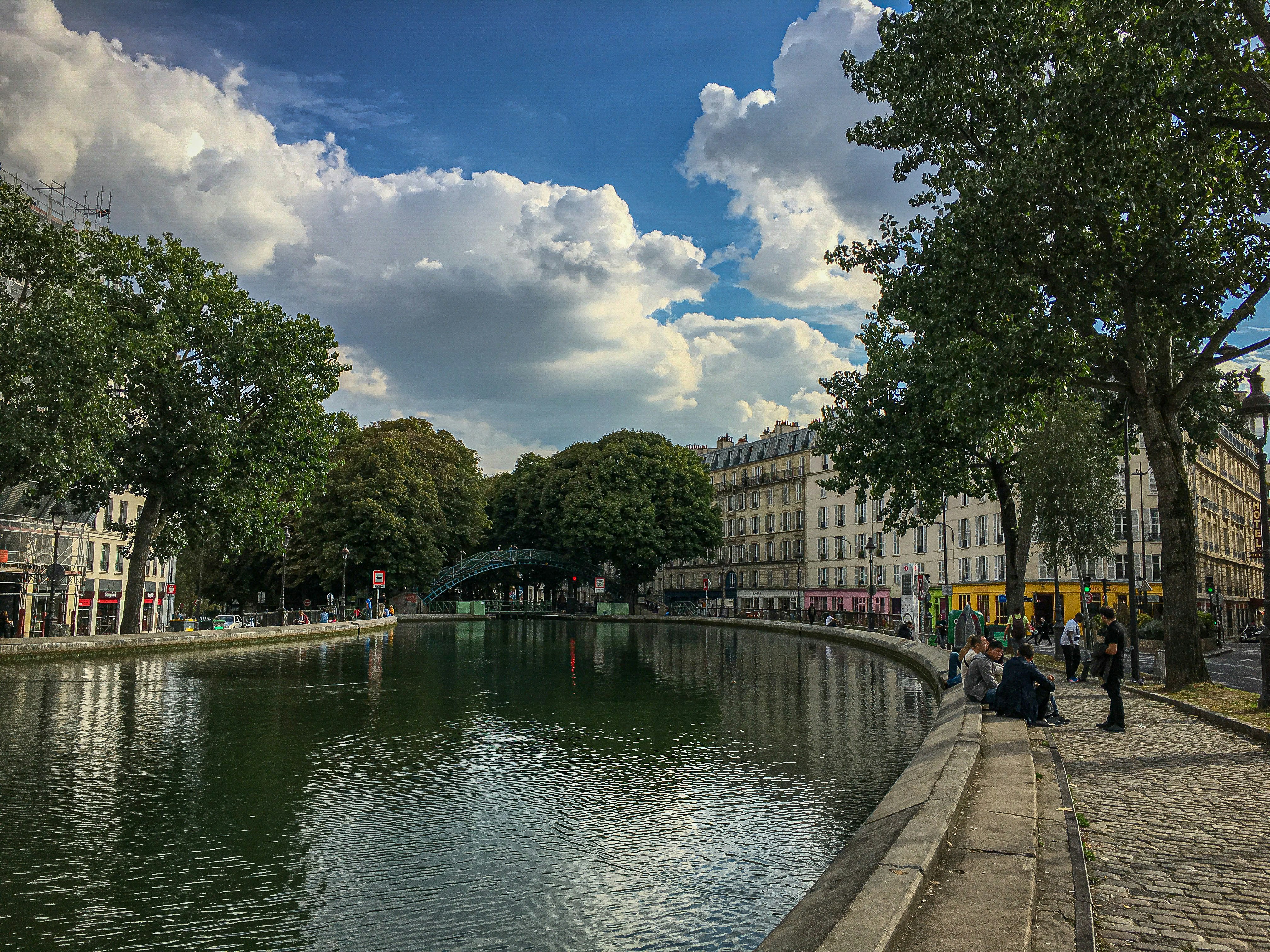 A calm canal flanked by trees and houses. People relax along the water.