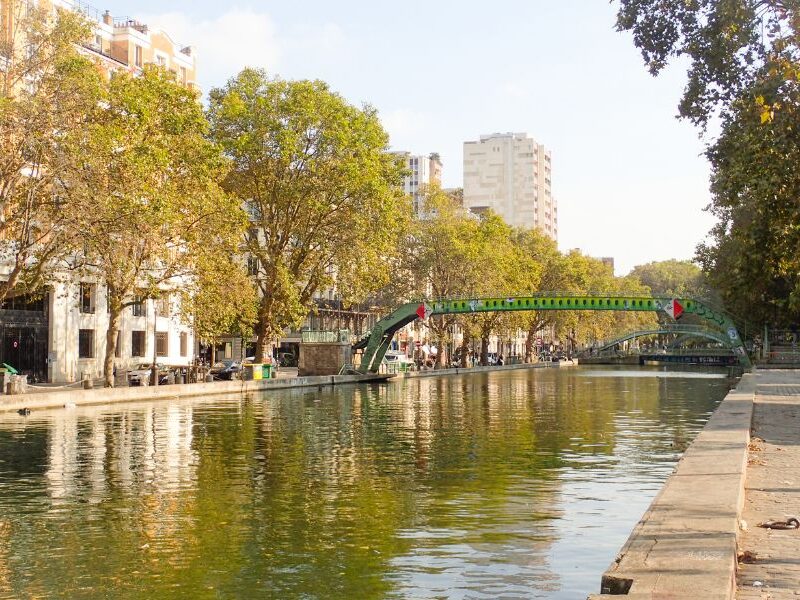 This is a picture of a canal in Paris. Over it is a small bridge and there are lots of green trees around.