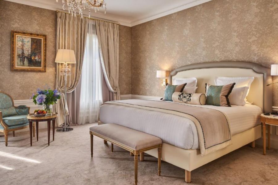 Grand hotel room in champagne and gold tones with damask wallpaper, crystal chandelier, Louis XVI-style furniture, teal accent cushions, and an ornate upholstered king bed