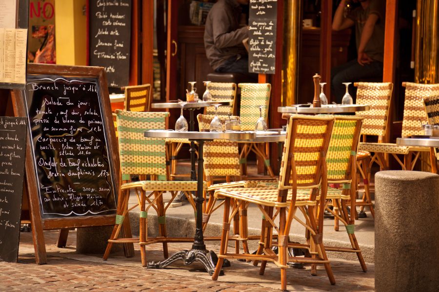 This is an image of the exterior of a cafe in Paris. It is lined with wooden tables and a big black board with a menu.