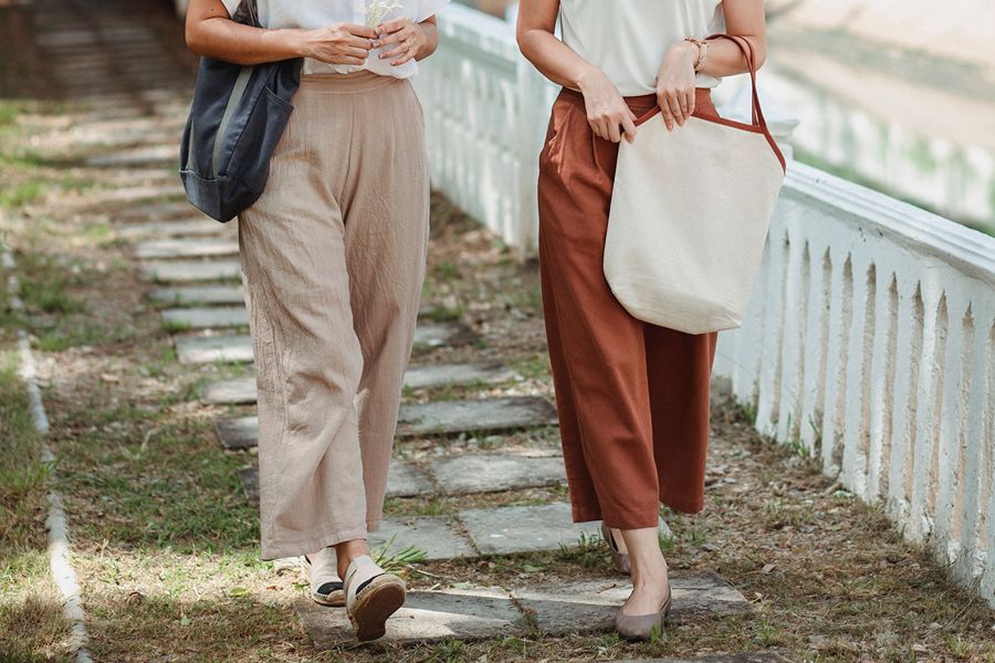 Close-up of two women's lower bodies walking along a garden path, one in beige linen wide-leg trousers with espadrilles, the other in rust-colored linen trousers with taupe flats, both carrying tote bags.