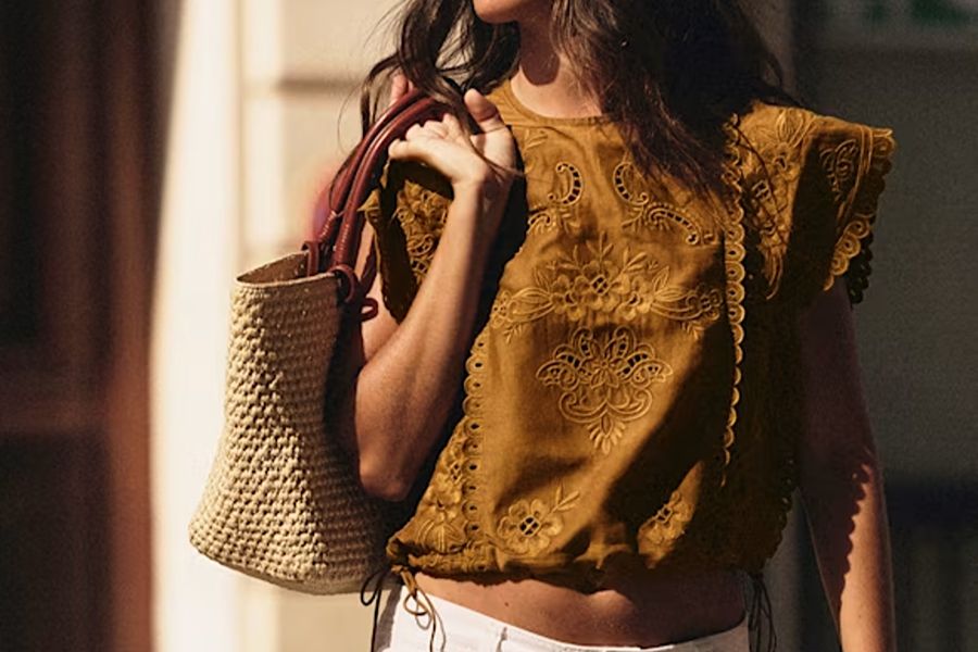 Close-up of a woman's torso wearing a mustard-yellow embroidered eyelet top with ruffled cap sleeves, carrying a woven raffia tote bag with burgundy leather handles.