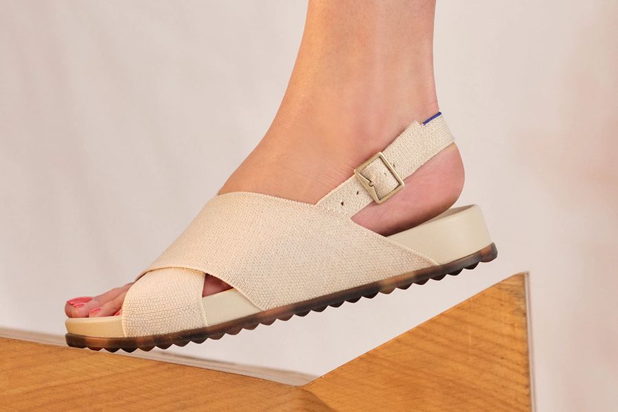 Close-up side view of a foot wearing a cream-colored woven crisscross-strap slingback sandal with a buckle, resting on a wooden ledge.