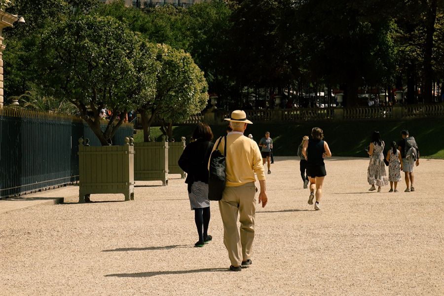 A man in a yellow sweater and beige trousers with a straw hat walks away down a gravel path in a Parisian park, with joggers and pedestrians in the background among trees.