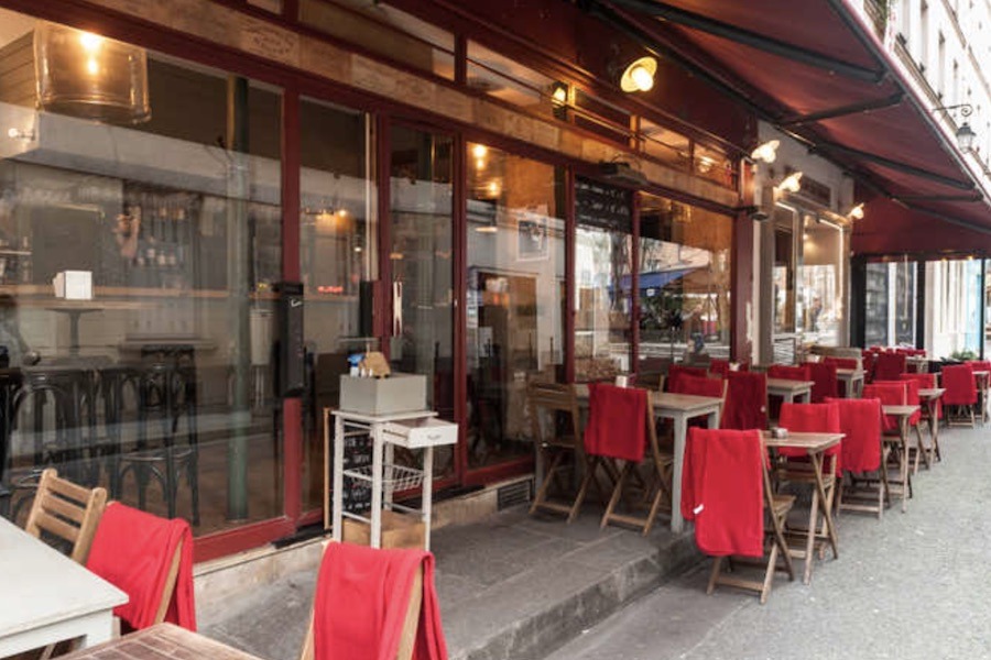 Exterior terrace of a classic Parisian wine bar with red-draped wooden chairs and small tables arranged on the pavement beneath a dark awning.