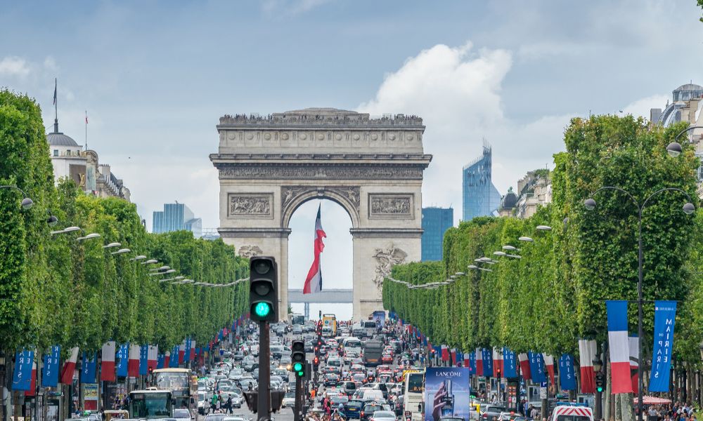 This is an image of the Champs Elysees with the Arc De Triomphe in the distance. The streets are full of cars and French flags.