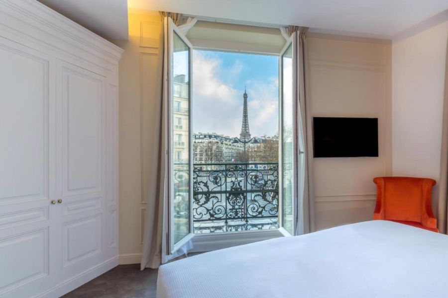 This is an image of a big, spacious hotel room. The bed has white bedding and there are big double doors that open up on to a traditional French balcony with the Eiffel Tower in the distance.