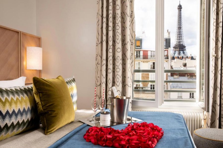 This is an image of a hotel bed with rose petals assembled into a heart and a bottle of champagne in an ice bucket. There is also a beautiful view out the window of the Eiffel Tower.
