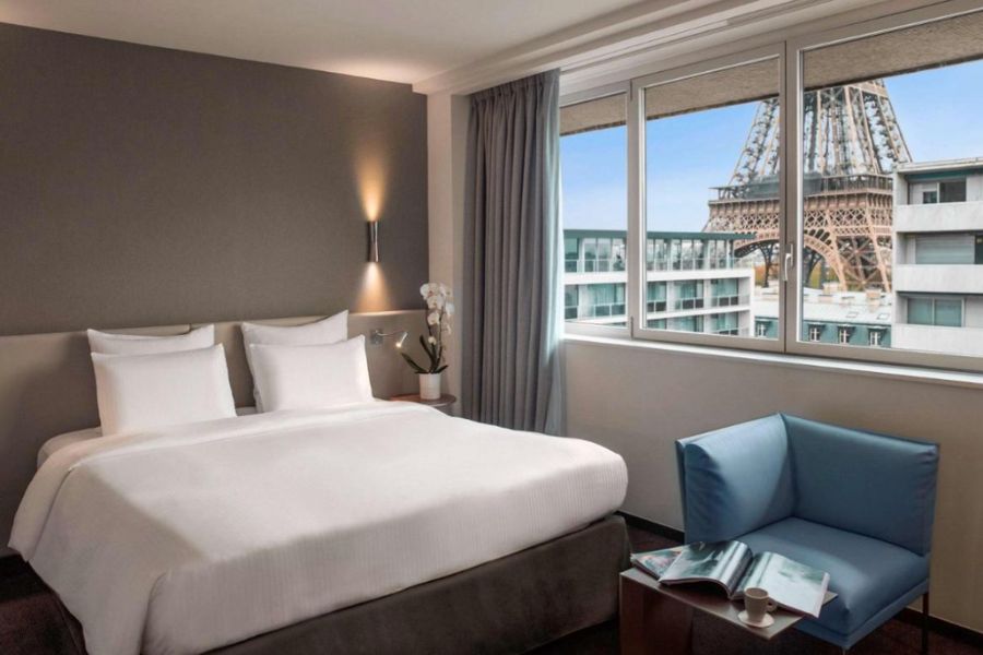 This is an image of a big, spacious hotel room with a large window where you can see the Eiffel Tower.