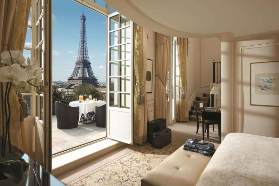 This is an image of a beautiful hotel room with a balcony you can walk out on. Out there, there are chairs, a table and a breakfast spread. The Eiffel Tower is in the distance.