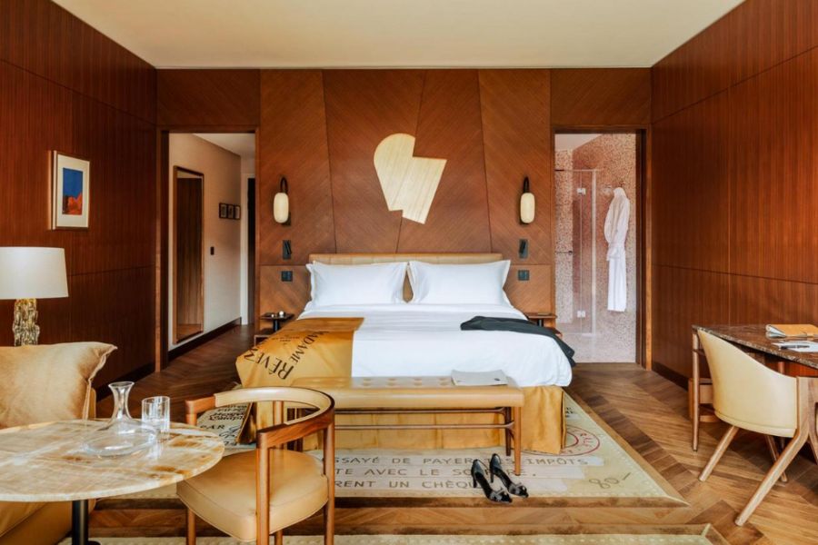 This is an image of a really spacious hotel room, with a bed in the centre and other amenities like a desk, dining table and rug. The decor and walls are a burnt orange contrasted with a white bed.
