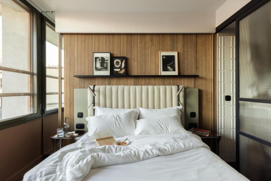 This is an image of a spacious hotel room with a bed in the centre, a wooden feature wall, a bedside table and some shelves with pictures on it.