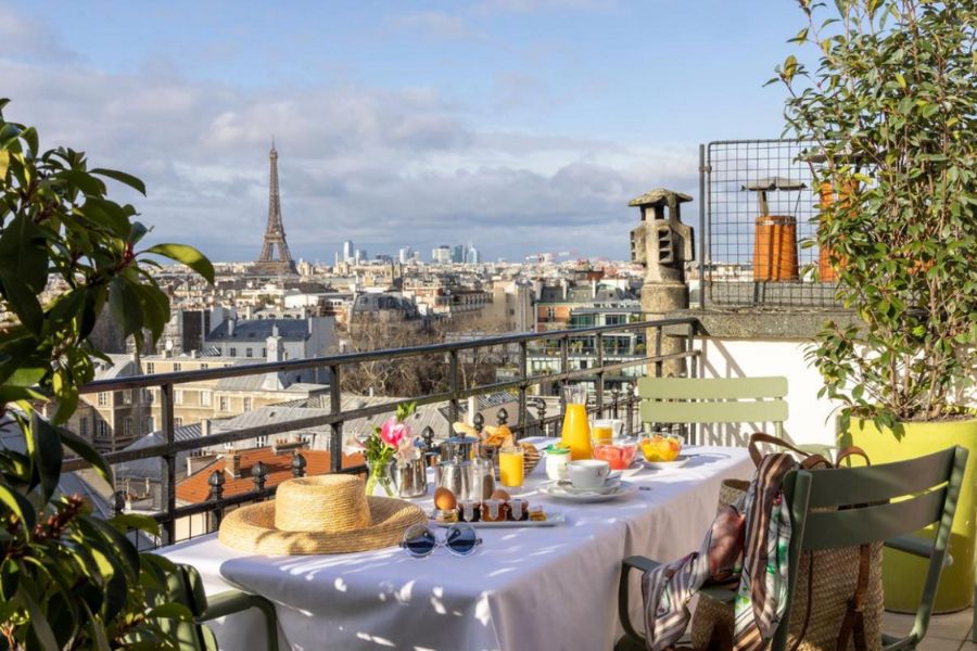 This is an image of a big balcony fit with a big table and breakfast spread, alongside a view of the Eiffel Tower in the distance.