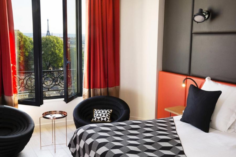This is an image of a red and black style room with a double bed and big windows that showcase a distant view of the Eiffel Tower.