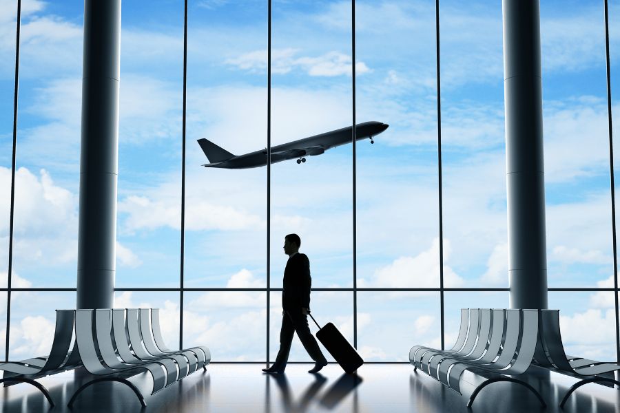 This is an image of a man walking in the airport terminal with his suitcase. There is a plane taking off in the background.
