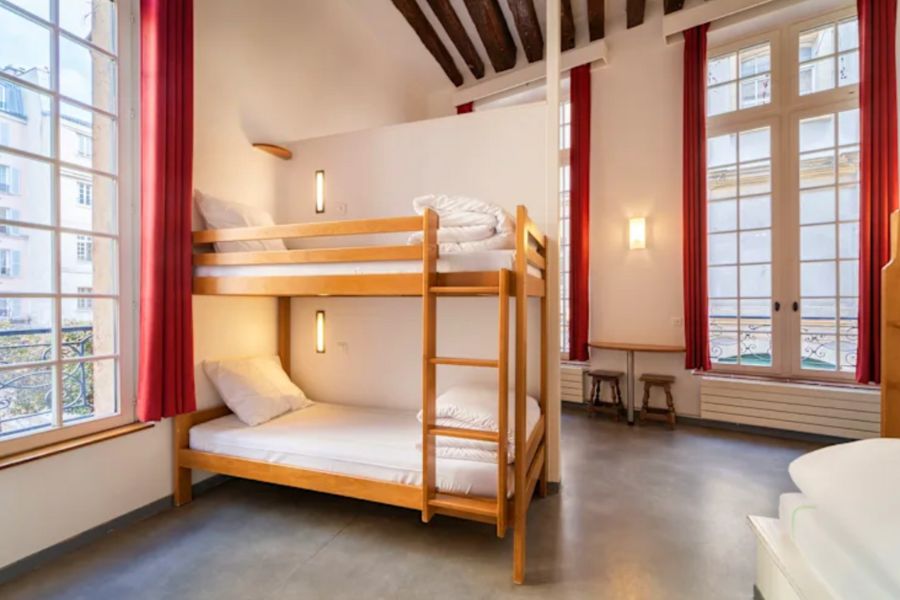 This is an image of a spacious hostel room with massive windows and several bunk beds.