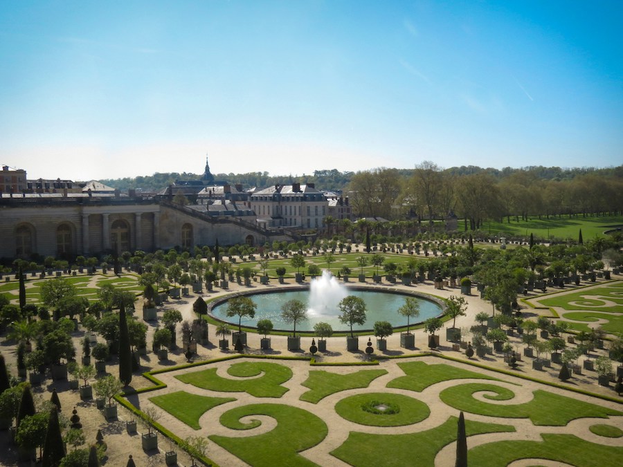 This is a picture of the gardens of Versailles. The plants and greenery are neatly gardened into swirls and regal shapes. The palace and water fountain can be seen in the centre.