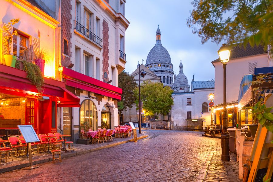 This is a picture of a well-lit Parisian street that has no tourists or people in it. Just a cafe with chairs. Areas like this are great to visit in Paris during the Olympics as they won't be as busy.