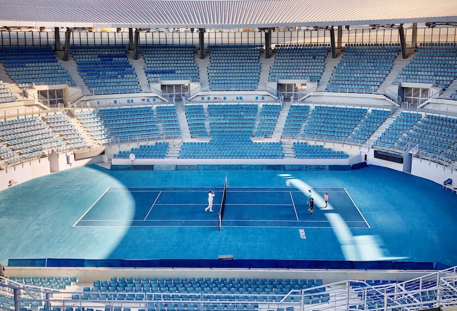 This is a picture of a massive arena that is not full yet. There is a tennis court with a few people playing in the centre.