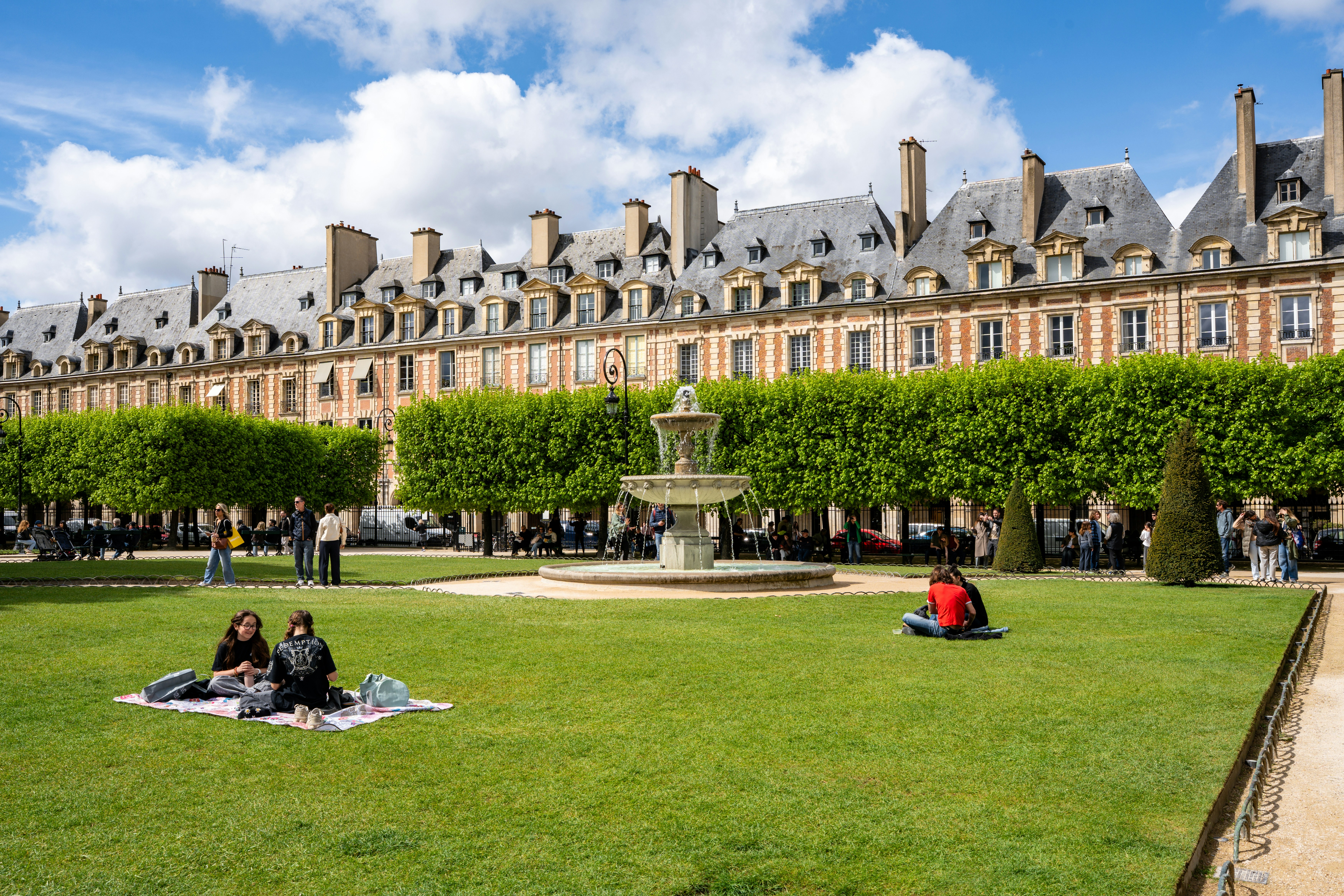 Red brick buildings surround a lush green square with trees and a fountain.